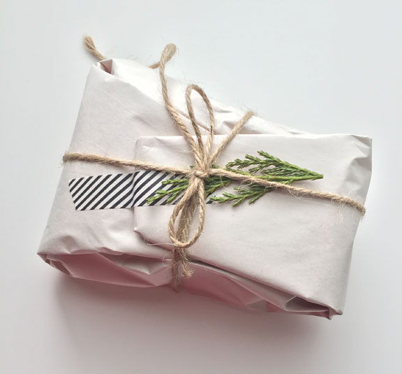 A package wrapped in white shipping paper and secured with black and white stripped washi tape. Tied with a string and adorned with a sprig of greenery