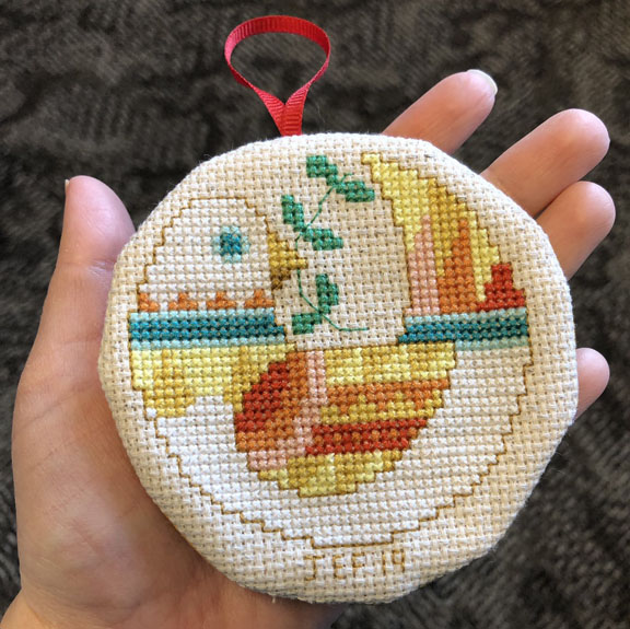 A person's hand holding a circular cross stitched ornament of a dove with an olive branch in its mouth.